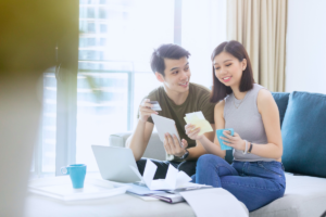 A couple sit on the couch in front there laptop discussing credit card debt. Both smiling, one is holding a credit card and one is holding bills.