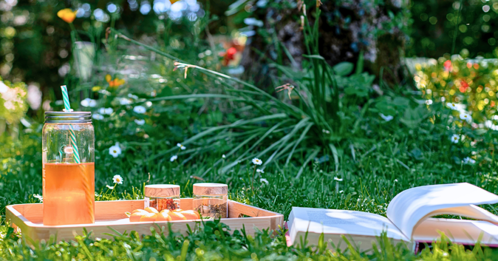 An open book and a tray with tea and snacks on a bed of grass shaded by a tree