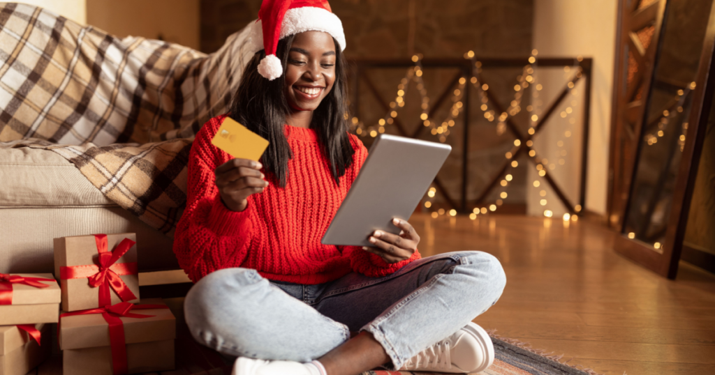 African American woman in holiday clothes holding a credit card and a tablet sitting on the floor by presents